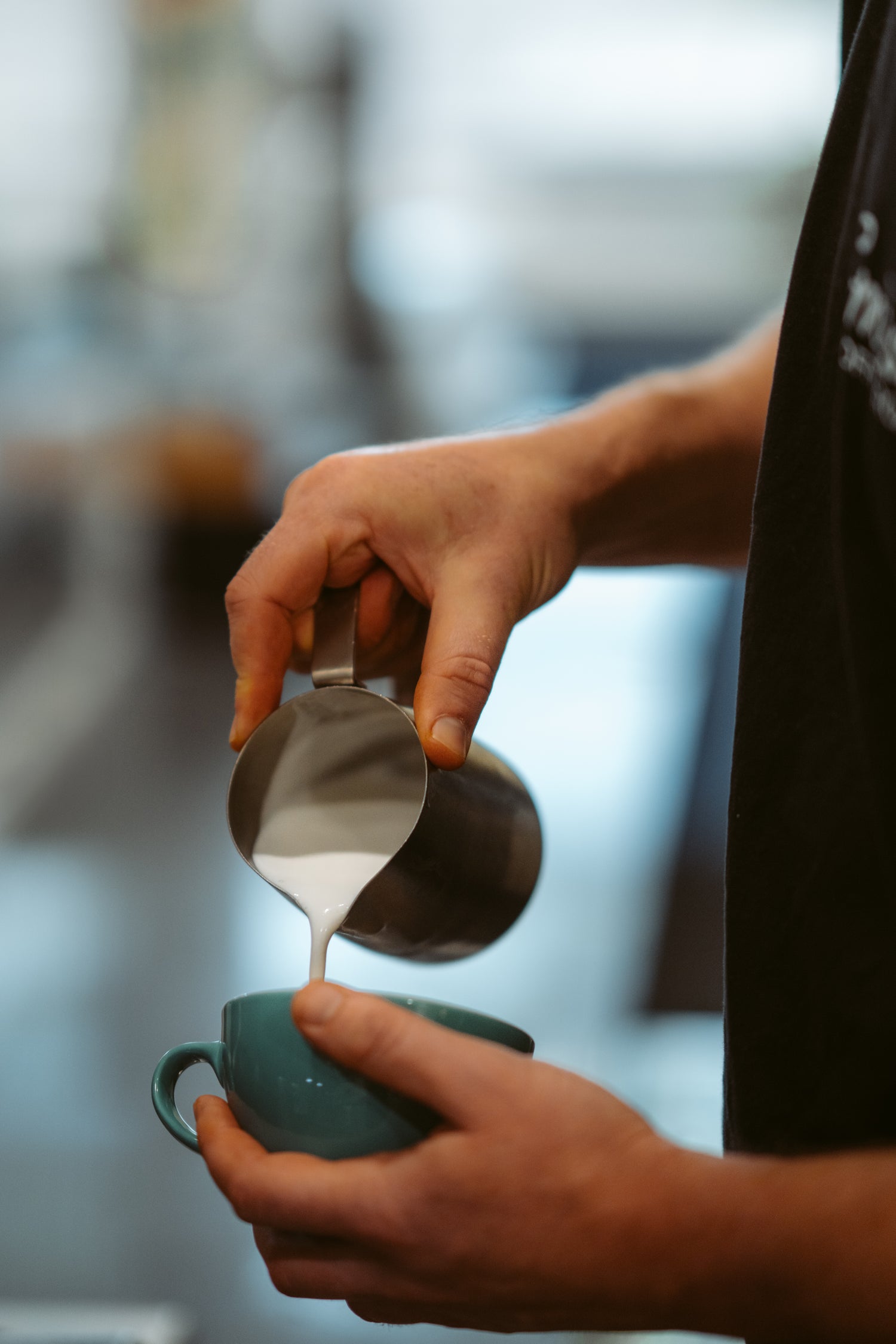 Person pouring steamed milk into a teal/blue cup with a blurred background