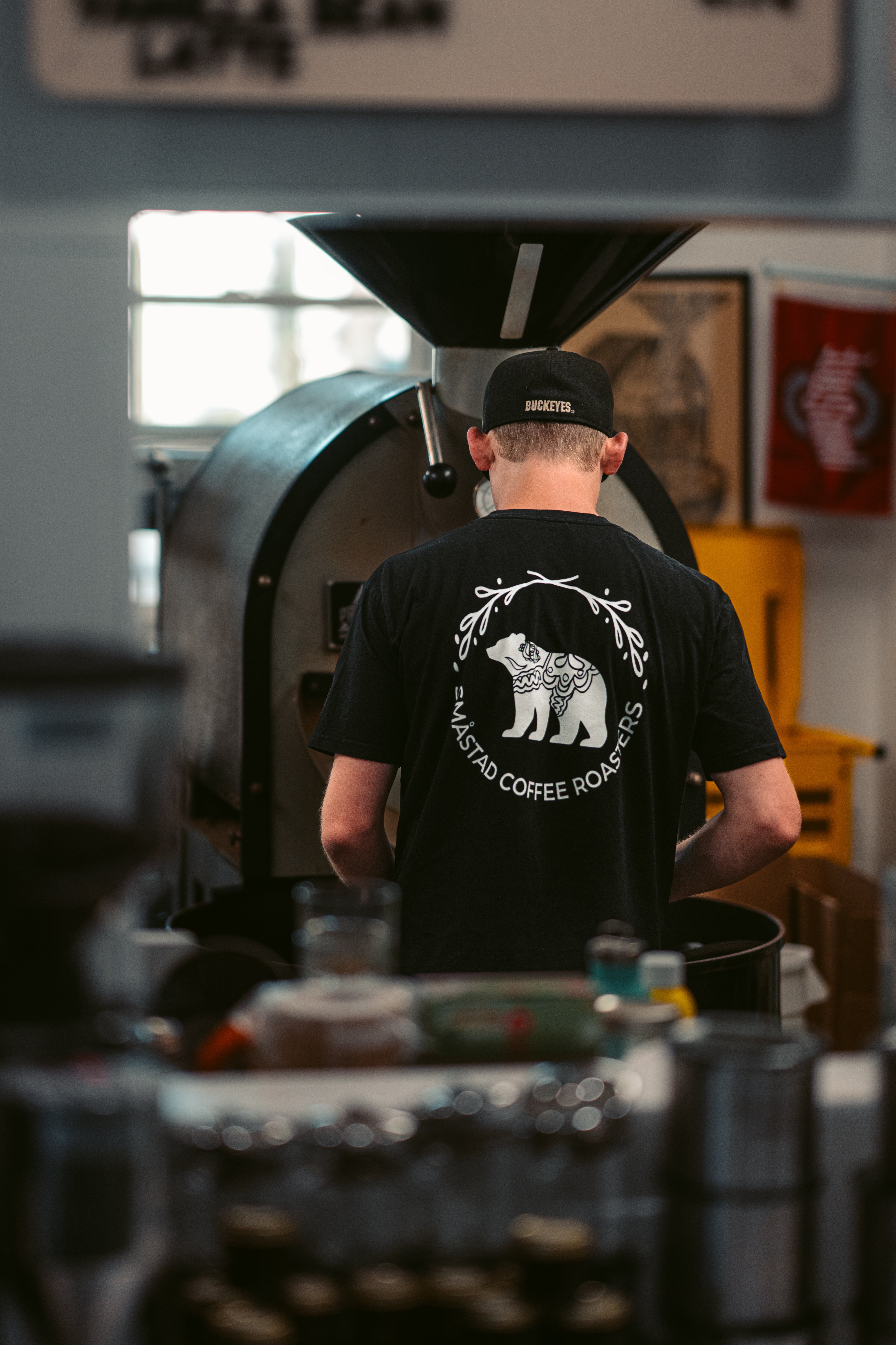 Person wearing a black t-shirt with a logo for Smastad Coffee Roasters in front of a coffee roasting machine.