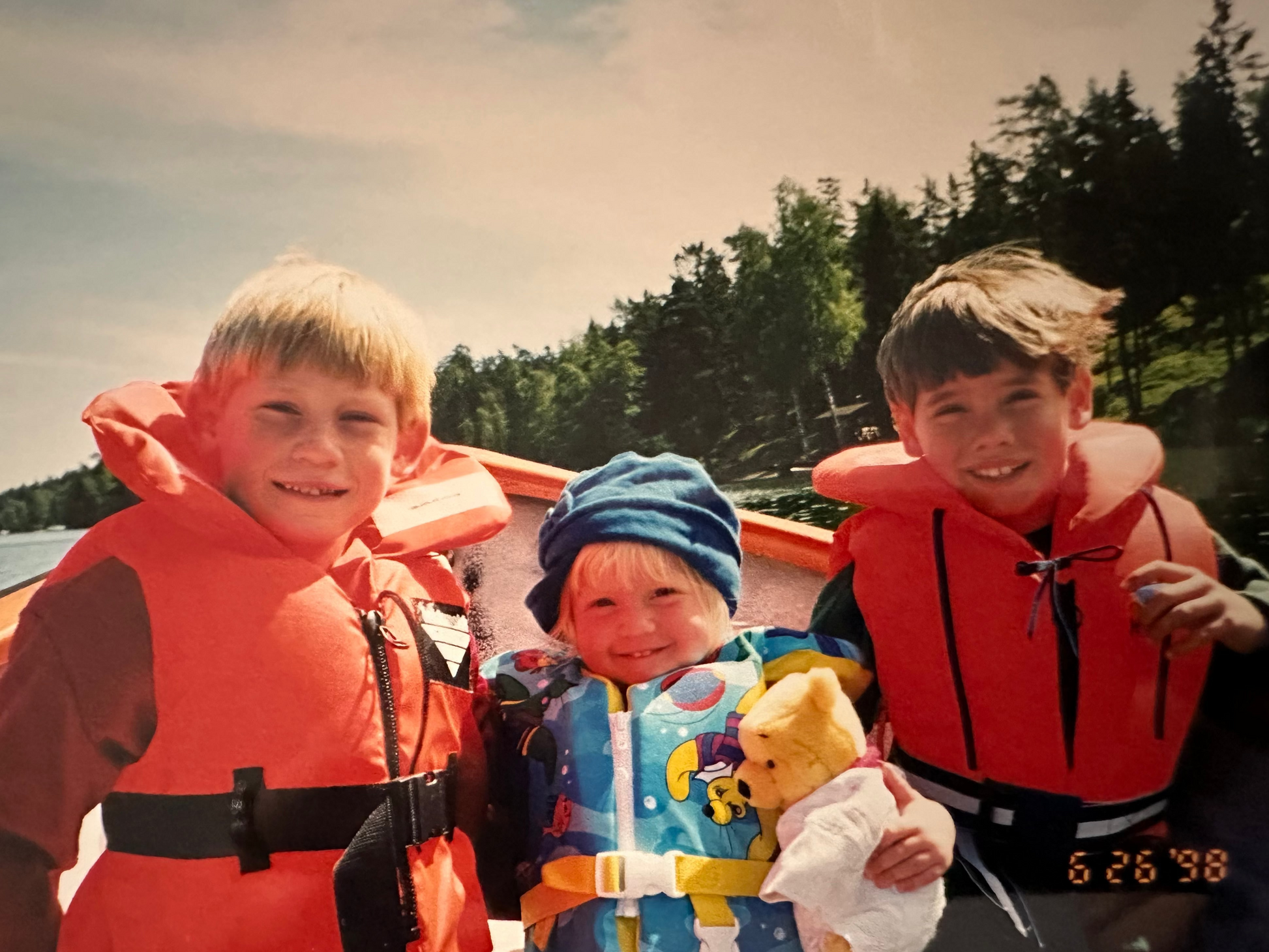 Three children in life jackets by a lake with trees in the background, taken on 6/26/98.